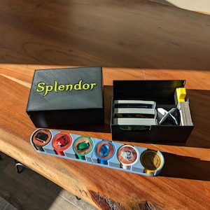 May include: A black Splendor game organiser with a lid, a card holder, and a token tray. The token tray holds various coloured tokens with gem and coin designs. The word "Splendor" is printed in gold on the lid.
