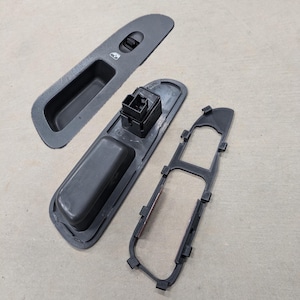 May include: A black plastic car door panel with a window switch and a door handle. The panel is disassembled into three pieces.