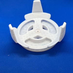 May include: White plastic base with three prongs and a circular centre with a small hole. The base is designed to hold a round object.