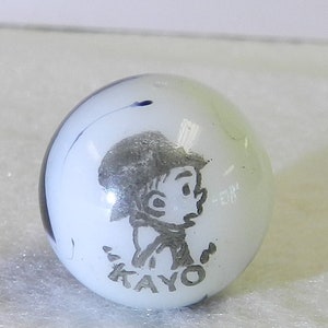 May include: White glass marble with a black and blue swirl design. The marble features a black and white image of a cartoon boy with the word "KAYO" printed below him.