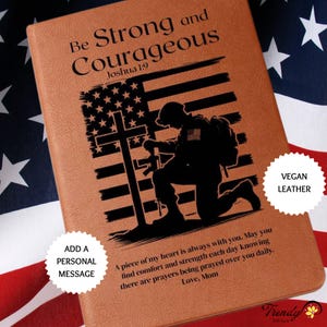 May include: A brown leather journal with a black silhouette of a soldier kneeling in prayer in front of a cross and an American flag. The text "Be Strong and Courageous" is at the top of the journal, with the scripture reference "Joshua 1:9" below. The words "Vegan Leather" are in a white circle on the right side of the journal. The text "Add a Personal Message" is in a white circle on the left side of the journal. The text "A piece of my heart is always with you. May you find comfort and strength each day knowing there are prayers being prayed over you daily. Love, Mom" is at the bottom of the journal.