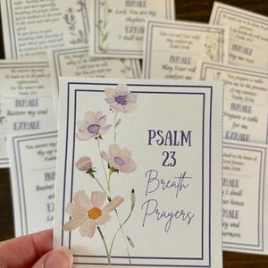 以下が含まれることがあります： 「Psalm 23 Breath Prayers」のテキストと花柄のデザインが施された正方形のカード。カードには、ピンクと紫の色合いの水彩画風の花が描かれており、青い縁取りがあります。テキストが書かれた他のカードが背景にあります。