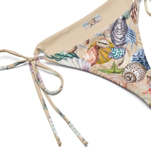 May include: A beige bikini bottom featuring a seashell print. The design includes various seashells, starfish, and coral in shades of beige, blue, pink, and green. The bikini has tie sides with a watercolor pattern.