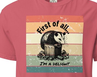May include: A vintage-style graphic t-shirt with a possum sitting in a trash can. The text reads "First of all... I'm a delight".