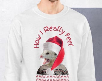 May include: White sweatshirt with a red and white possum wearing a Santa hat. The possum is wearing a sweater with a pattern of red and white stripes. The text "How I Really Feel" is written in red around the possum. The text "AAAAAAA AAAAAAAA AAAAAAAA AAAAAAAA AAAAAAH" is written in red below the possum.