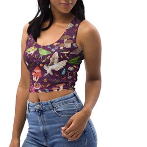 May include: A sleeveless crop top in a deep purple hue, adorned with a whimsical pattern of mushrooms, insects, and an owl. The design features a scoop neckline and is made of a soft, comfortable material. The top is paired with blue denim jeans.