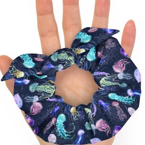 Peut inclure: Un chouchou bleu foncé avec un motif de méduses colorées sur fond noir. Le chouchou est noué et est tenu dans une main.