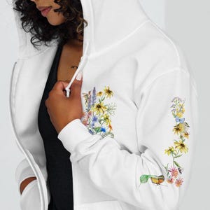 May include: White zippered hoodie with a floral design on the sleeve. The design features a variety of flowers, leaves, and a small bird.