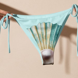 May include: Light blue bikini bottom with tie sides. The front features a seashell design in shades of teal, beige, and white, with a pearl accent at the bottom. The bikini is held up by hands.