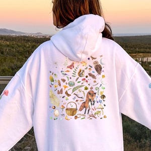 May include: White hoodie with a colorful print of various woodland creatures, including a fox, owl, squirrel, and mushrooms.