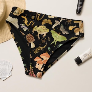 May include: Black high-waisted bikini bottoms with a whimsical pattern of mushrooms, insects, frogs, and butterflies. The design features a variety of colours, including green, orange, and yellow, set against a black background. The bottoms are made of a soft, comfortable material.