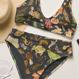 May include: Black bikini with a dark floral print featuring butterflies, mushrooms, frogs, and other woodland creatures. The print is on a dark background with gold glitter accents.