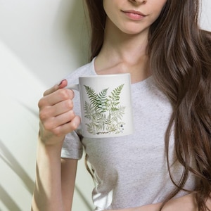 May include: White ceramic mug with a green fern design. The mug has a handle and is filled with a white liquid.