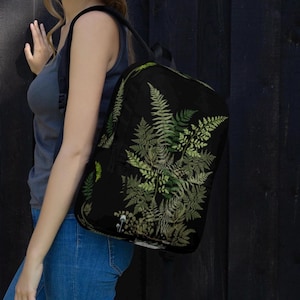 May include: Black backpack featuring a detailed green fern design. The backpack has a black shoulder strap and a black zipper. The fern design is a mix of light and dark green, creating a natural, botanical aesthetic. The backpack is made of a durable material.