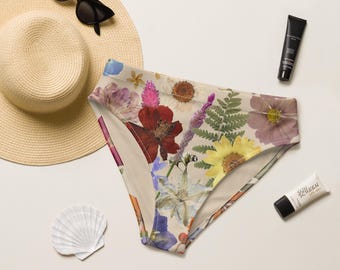 May include: High-waisted bikini bottoms featuring a colorful pressed flower and fern print, accompanied by a straw sun hat, black sunglasses, a tube of sunscreen, and a white seashell.