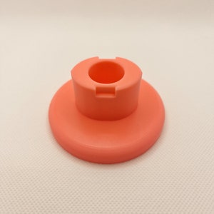 May include: A coral-coloured plastic stand with a round base and a raised centre ring. The stand is designed to hold a small object, such as a phone or a candle.