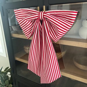 May include: A large decorative bow made of red and white striped fabric. The bow is tied in the center and hangs from a black cabinet with glass doors. The stripes are vertical and evenly spaced.