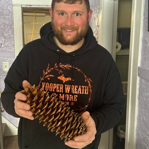 Op de afbeelding: Een persoon houdt een grote, bruine dennenappel vast. De dennenappel is ongeveer 30 cm lang en heeft een ruw, getextureerd oppervlak. De persoon draagt een zwarte hoodie met de tekst "Yooper Wreath & More" erop gedrukt. De tekst is koperkleurig.