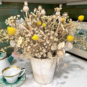 May include: A dried flower arrangement in a weathered, off-white pot. The arrangement features beige, yellow, and tan dried flowers, including seed pods and small, round yellow blooms. A teacup and saucer with gold trim and a green geometric design are in the foreground.