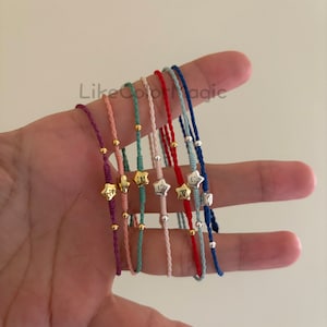 May include: A set of eight colourful friendship bracelets with a gold star charm. Each bracelet is made with a different colour of string and has a small gold bead on each end.
