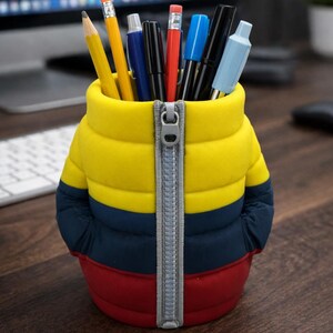 May include: A desk organizer shaped like a puffer jacket, in yellow, navy blue, and red. The jacket-shaped container holds various pens and pencils. The zipper is silver. The desk organizer is on a wooden desk.