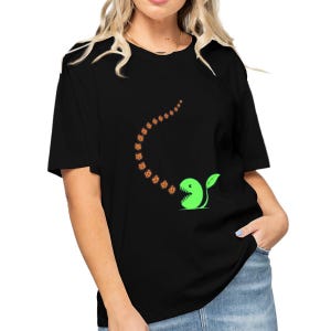 Puede incluir: Camiseta negra con un diseño de personaje de dibujos animados verde. El personaje tiene una hoja y la boca abierta, emitiendo un rastro de formas naranjas. El diseño está centrado en la parte delantera. La camiseta tiene mangas cortas y cuello redondo.