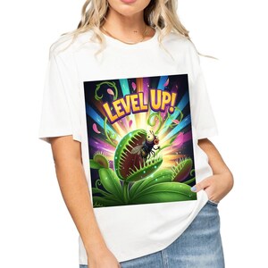 Puede incluir: Camiseta blanca con un gráfico colorido que dice "LEVEL UP!". El gráfico representa una Venus atrapamoscas con una mosca dentro. El diseño tiene un estilo vibrante de dibujos animados con colores brillantes y un tema de juego.