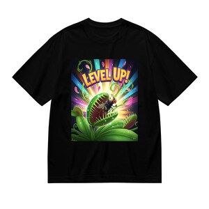 Puede incluir: Camiseta negra con un gráfico colorido de una Venus atrapamoscas con una mosca y la frase "LEVEL UP!". El diseño tiene un estilo vibrante y de dibujos animados con colores brillantes y un tema lúdico.