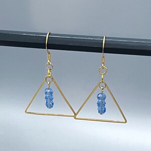 May include: Gold-tone triangle earrings with three light blue beads dangling from the center. The earrings have a simple, geometric design and are suspended from gold-colored ear wires.