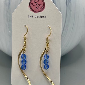May include: Gold dangle earrings with three light blue faceted beads hanging from a curved wire. The earrings are on a white card with the text "SHE Designs" printed on it.