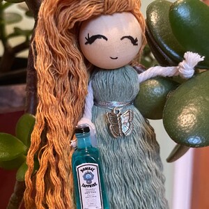 May include: A doll with long, orange hair is holding a miniature bottle of Bombay Sapphire gin. The doll is wearing a light blue dress with a silver butterfly charm.