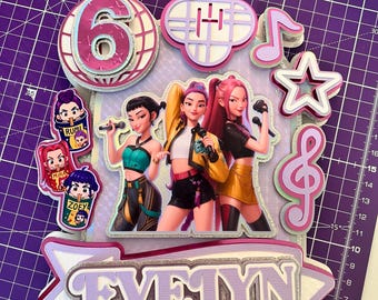 May include: A layered cake topper with a music theme. It features a number 6, musical notes, stars, and a banner that reads "EVELYN". The design includes cartoon figures of singers and the names "RUMI" and "ZOEY". The color scheme is primarily pink, purple, and silver.