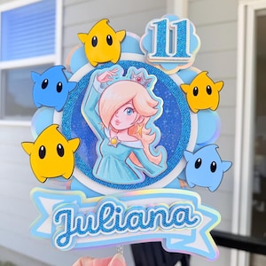 May include: A layered paper cake topper featuring a central illustration of a blonde character in a blue dress, surrounded by yellow and blue star-shaped figures. The topper includes the number 11 in blue glitter and the name Juliana in blue script.