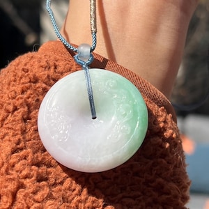 May include: A jade pendant necklace with a round, green and white jade pendant. The pendant has intricate carvings and is suspended from a blue cord.