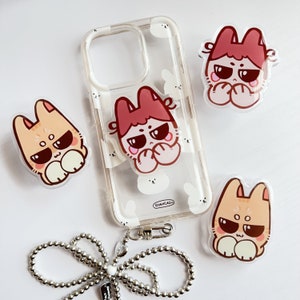 May include: A clear phone case with a white and pink design featuring cartoon characters. The case has a ring holder with a pink and red cartoon character. A silver chain is attached to the case. There are two additional pink and red cartoon character phone grips.