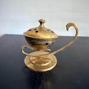 May include: A vintage brass incense burner with a decorative lid and a curved handle. The burner has a round base and a bowl-shaped body with floral cutouts. The brass has a warm, golden hue, and the design suggests an antique or ornamental piece.