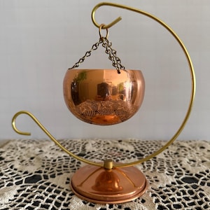 May include: A copper incense burner with an etched design, suspended by a chain from a curved brass stand. The stand has a circular base. The burner is designed to hold incense.