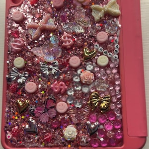 May include: Pink tablet case decorated with glitter, bows, butterflies, smiley faces, and heart-shaped charms. The case has a variety of embellishments in shades of pink, white, and gold. The word "GURL" is spelled out in letter beads.