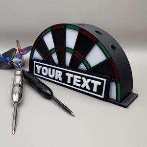 May include: A black and white dartboard-shaped holder with the text "YOUR TEXT" on a white rectangle. Three darts are next to the holder. The dartboard has red and green accents.