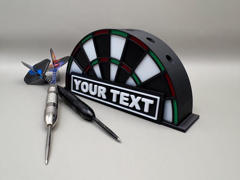 May include: A black and white dartboard-shaped holder with the text "YOUR TEXT" on a white rectangle. Three darts are next to the holder. The dartboard has red and green accents.