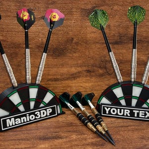 May include: A set of darts with black, silver, and colorful flights, displayed in a black and white dartboard-shaped holder. One holder reads "Manio3DP" and the other reads "YOUR TEXT". The darts are on a wooden surface.