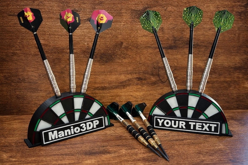 May include: A set of darts with black, silver, and colorful flights, displayed in a black and white dartboard-shaped holder. One holder reads "Manio3DP" and the other reads "YOUR TEXT". The darts are on a wooden surface.