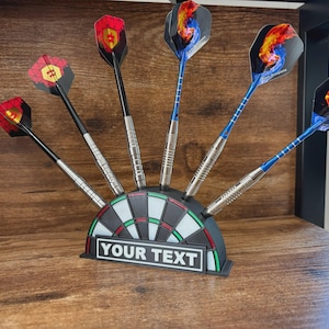 May include: A set of darts with colorful flights, displayed in a dartboard-shaped holder. The darts have black, silver, and blue shafts, with various flame and geometric designs on the flights. The holder has the text "YOUR TEXT".