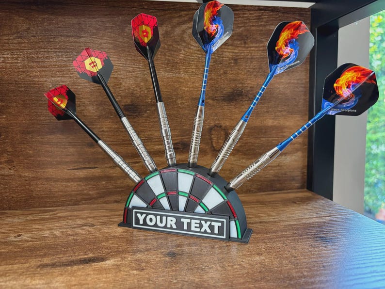 May include: A set of darts with colorful flights, displayed in a dartboard-shaped holder. The darts have black, silver, and blue shafts, with various flame and geometric designs on the flights. The holder has the text "YOUR TEXT".