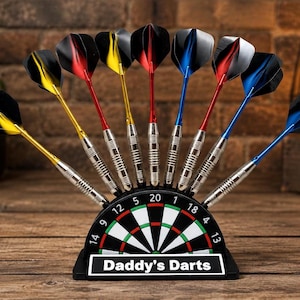 May include: A collection of darts with black, yellow, red, and blue flights, displayed in a black dartboard-shaped holder. The holder is inscribed with "Daddy's Darts". The darts are presented on a wooden surface.