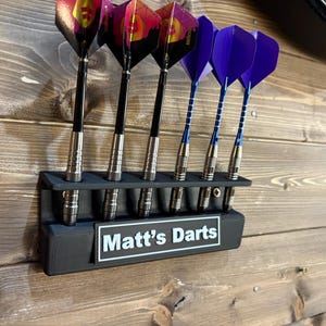 Może przedstawiać: Czarny uchwyt na lotki z sześcioma miejscami na lotki. Uchwyt ma białą etykietę z napisem "Matt's Darts". W uchwycie znajduje się sześć lotek, trzy z fioletowymi piórami i trzy z czerwonymi i czarnymi piórami.
