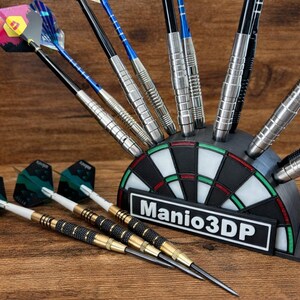 May include: A black and white dartboard-shaped holder with darts. The holder has the text "Manio3DP" on it. Several darts are in the holder, and some are lying on the wooden surface. The darts have various colors and designs.