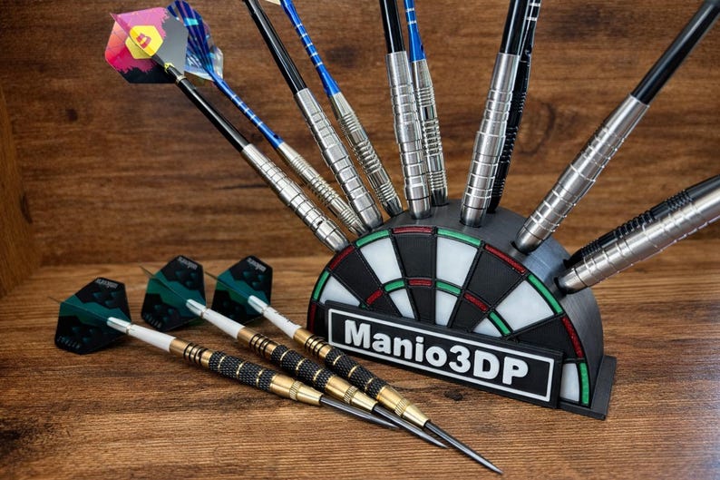 May include: A black and white dartboard-shaped holder with darts. The holder has the text "Manio3DP" on it. Several darts are in the holder, and some are lying on the wooden surface. The darts have various colors and designs.