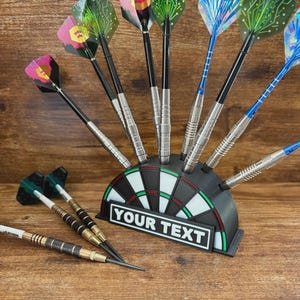 May include: A collection of darts with various colorful flights, displayed in a black dartboard-shaped holder. The holder has a white rectangle with the text "YOUR TEXT". The darts have silver and black shafts. Some darts are lying on a wooden surface.
