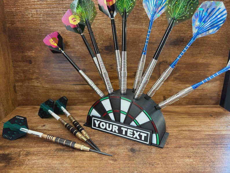 May include: A collection of darts with various colorful flights, displayed in a black dartboard-shaped holder. The holder has a white rectangle with the text "YOUR TEXT". The darts have silver and black shafts. Some darts are lying on a wooden surface.